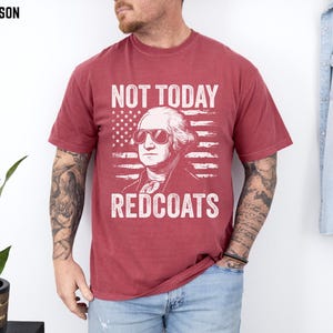 May include: Crimson t-shirt with a graphic of George Washington in sunglasses, set against an American flag. The text reads "NOT TODAY REDCOATS" in white. A casual wear item, suitable for everyday use.