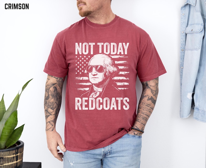 May include: Crimson t-shirt with a graphic of George Washington in sunglasses, set against an American flag. The text reads "NOT TODAY REDCOATS" in white. A casual wear item, suitable for everyday use.