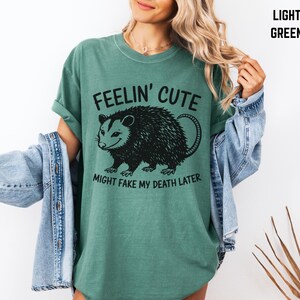 Może przedstawiać: Jasnozielona koszulka z napisem "Feelin' Cute Might Fake My Death Later" i czarną grafiką oposa. Koszulka ma luźny kr&oacute;j i podwinięte rękawy. Osoba nosi jasnoniebieską kurtkę dżinsową.