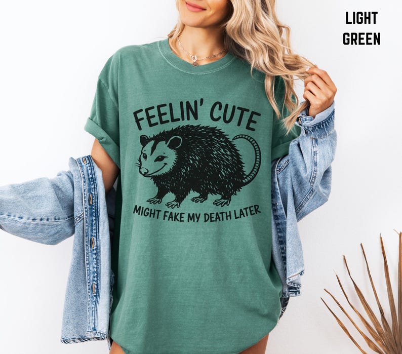 Może przedstawiać: Jasnozielona koszulka z napisem "Feelin' Cute Might Fake My Death Later" i czarną grafiką oposa. Koszulka ma luźny kr&oacute;j i podwinięte rękawy. Osoba nosi jasnoniebieską kurtkę dżinsową.