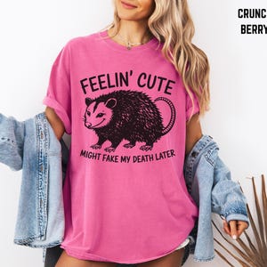 Może przedstawiać: R&oacute;żowa koszulka z napisem "FEELIN' CUTE" nad czarną ilustracją oposa i frazą "MIGHT FAKE MY DEATH LATER". Koszulka jest w jednolitym kolorze i ma kr&oacute;tkie rękawy.