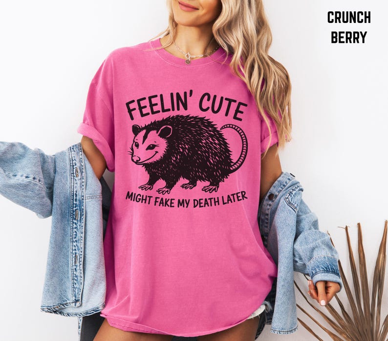 Może przedstawiać: R&oacute;żowa koszulka z napisem "FEELIN' CUTE" nad czarną ilustracją oposa i frazą "MIGHT FAKE MY DEATH LATER". Koszulka jest w jednolitym kolorze i ma kr&oacute;tkie rękawy.
