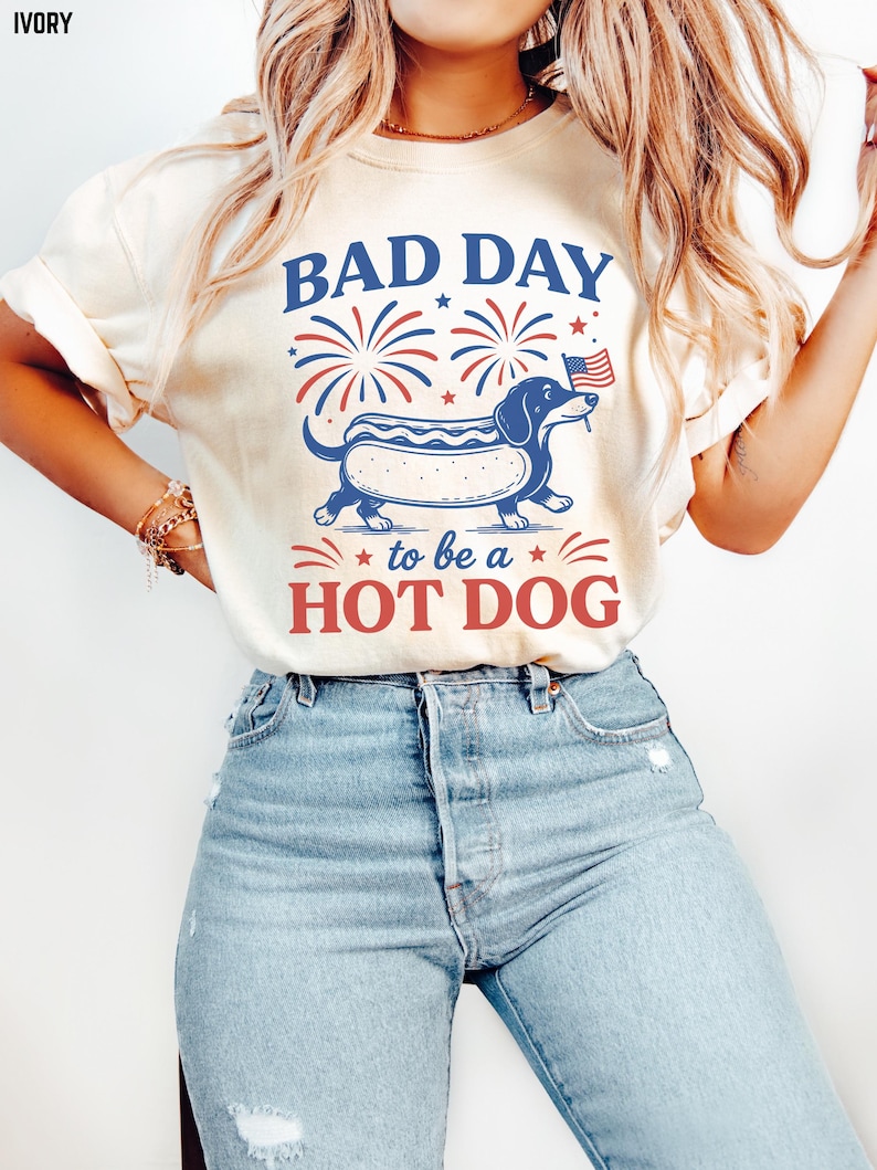 May include: Ivory-colored t-shirt with the text "BAD DAY to be a HOT DOG" in red and blue. The graphic features a dachshund hot dog with an American flag. Fireworks and stars complete the patriotic design.