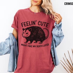 Może przedstawiać: Koszulka w kolorze karmazynowym z napisem "FEELIN' CUTE" nad czarną ilustracją oposa i napisem "MIGHT FAKE MY DEATH LATER". Koszulka ma kr&oacute;tkie rękawy i luźny kr&oacute;j.