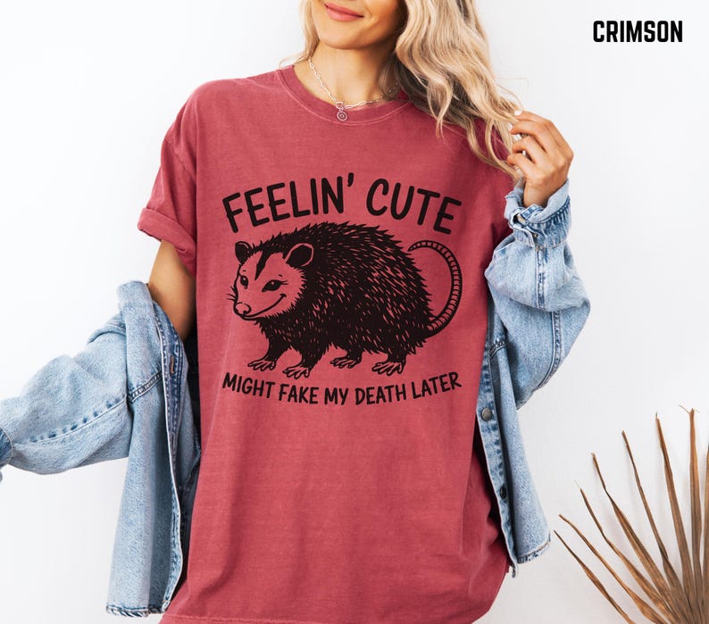 Może przedstawiać: Koszulka w kolorze karmazynowym z napisem "FEELIN' CUTE" nad czarną ilustracją oposa i napisem "MIGHT FAKE MY DEATH LATER". Koszulka ma kr&oacute;tkie rękawy i luźny kr&oacute;j.