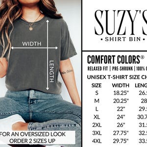 May include: A dark grey unisex t-shirt with a size chart and the text "SUZY'S SHIRT BIN." The shirt is shown on a person wearing blue jeans. The size chart shows measurements in inches for width and length.