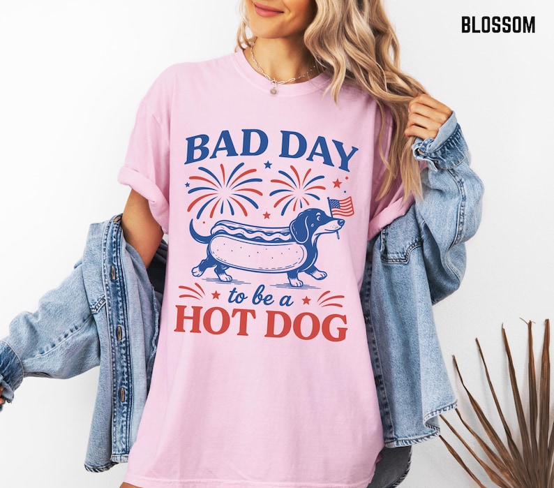 May include: A light pink t-shirt with the text "BAD DAY to be a HOT DOG" in blue and red. The graphic features a dachshund hot dog with an American flag. The shirt has rolled-up sleeves.