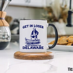 Pu&ograve; includere: Tazza in ceramica bianca con manico e bordo neri. La tazza presenta il testo "GET IN LOSER" sopra un'illustrazione di un uomo in barca, con il testo "WE'RE CROSSING THE DELAWARE" sotto. La tazza &egrave; di 312 grammi.