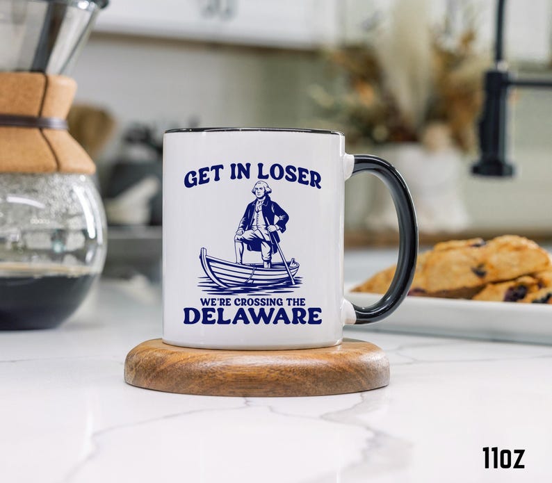 Pu&ograve; includere: Tazza in ceramica bianca con manico e bordo neri. La tazza presenta il testo "GET IN LOSER" sopra un'illustrazione di un uomo in barca, con il testo "WE'RE CROSSING THE DELAWARE" sotto. La tazza &egrave; di 312 grammi.