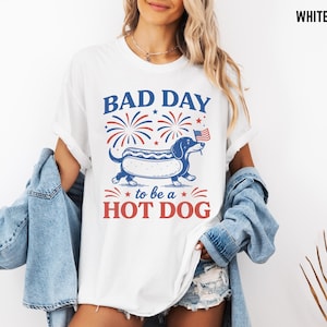 May include: White t-shirt with a graphic of a dachshund hot dog, fireworks, and the text "BAD DAY to be a HOT DOG" in red and blue. The dachshund is blue with a hot dog bun body and holding a small American flag.