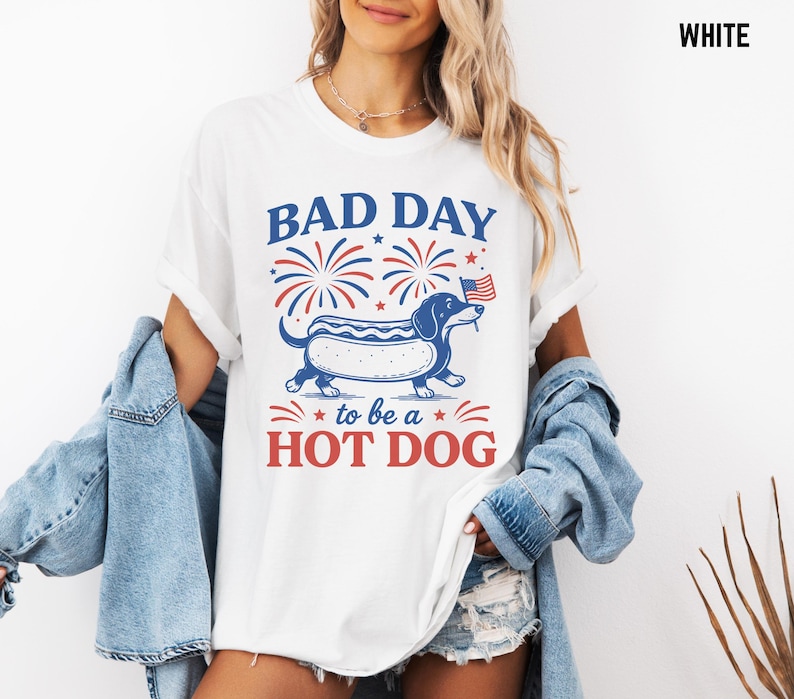 May include: White t-shirt with a graphic of a dachshund hot dog, fireworks, and the text "BAD DAY to be a HOT DOG" in red and blue. The dachshund is blue with a hot dog bun body and holding a small American flag.