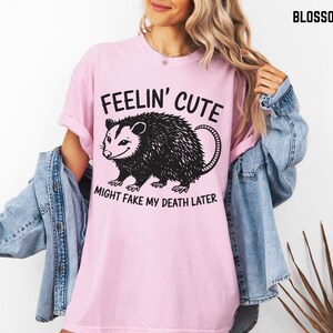 Może przedstawiać: Jasnor&oacute;żowa koszulka z napisem "Feelin' Cute Might Fake My Death Later" i czarną grafiką oposa. Koszulka ma kr&oacute;tkie rękawy i luźny kr&oacute;j.