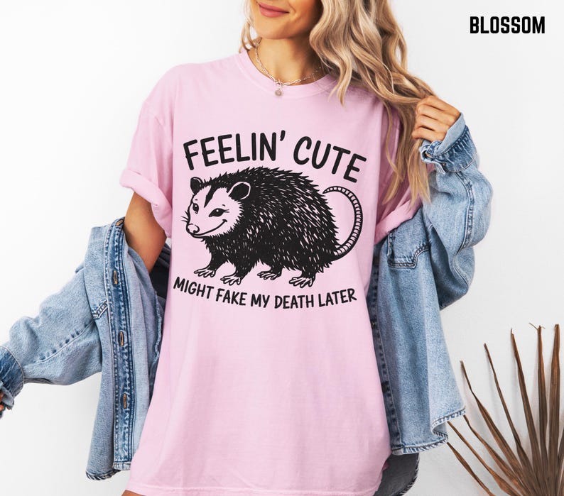 Może przedstawiać: Jasnor&oacute;żowa koszulka z napisem "Feelin' Cute Might Fake My Death Later" i czarną grafiką oposa. Koszulka ma kr&oacute;tkie rękawy i luźny kr&oacute;j.