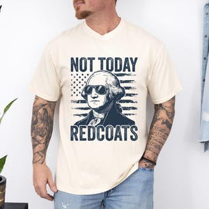 May include: Ivory t-shirt with a graphic of George Washington in sunglasses, and the text "NOT TODAY REDCOATS". The design includes an American flag. The shirt is a crew neck. The t-shirt is a cream colour.