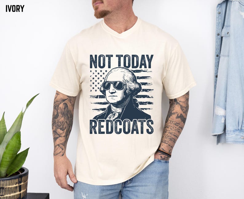 May include: Ivory t-shirt with a graphic of George Washington in sunglasses, and the text "NOT TODAY REDCOATS". The design includes an American flag. The shirt is a crew neck. The t-shirt is a cream colour.