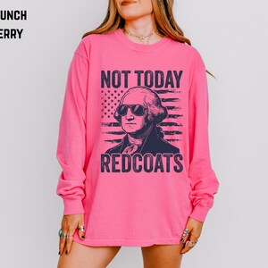 May include: A long-sleeved, bright pink shirt with a graphic of George Washington wearing sunglasses. The text "NOT TODAY REDCOATS" is printed above and below the image of the American flag.