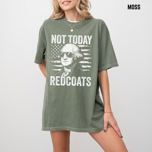 May include: Moss green t-shirt with a graphic of George Washington in sunglasses and an American flag design. The text reads "NOT TODAY REDCOATS". The shirt has a relaxed fit. The colour is a muted green.