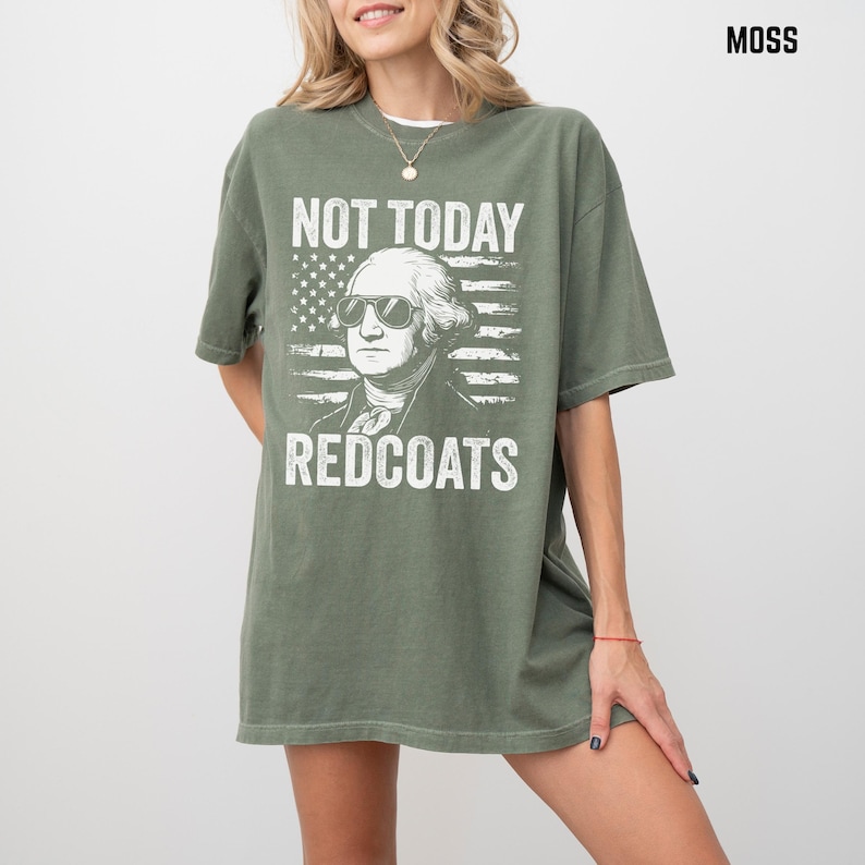 May include: Moss green t-shirt with a graphic of George Washington in sunglasses and an American flag design. The text reads "NOT TODAY REDCOATS". The shirt has a relaxed fit. The colour is a muted green.