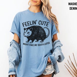 Może przedstawiać: Jasnoniebieska koszulka z czarną grafiką oposa i napisem "FEELIN' CUTE MIGHT FAKE MY DEATH LATER". Koszulka ma kr&oacute;tkie rękawy i jest wykonana z miękkiego materiału.