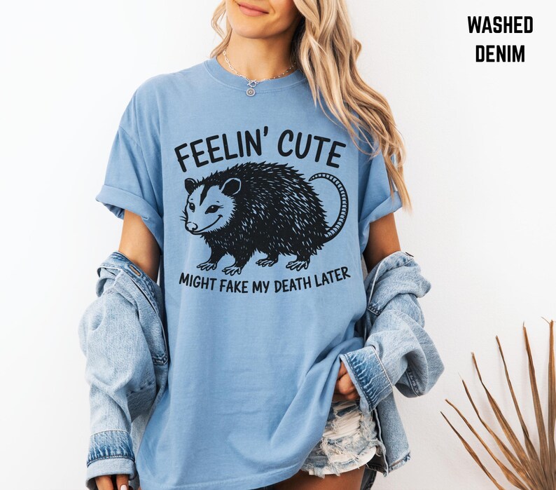 Może przedstawiać: Jasnoniebieska koszulka z czarną grafiką oposa i napisem "FEELIN' CUTE MIGHT FAKE MY DEATH LATER". Koszulka ma kr&oacute;tkie rękawy i jest wykonana z miękkiego materiału.