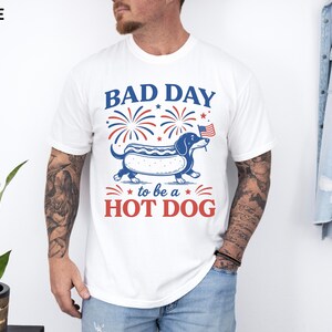 May include: White t-shirt with a graphic of a dachshund in a hot dog bun, holding a small American flag. The text reads "BAD DAY to be a HOT DOG" in red and blue. Fireworks are above the dog.