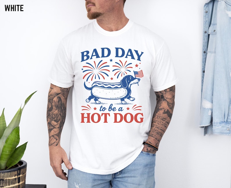 May include: White t-shirt with a graphic of a dachshund in a hot dog bun, holding a small American flag. The text reads "BAD DAY to be a HOT DOG" in red and blue. Fireworks are above the dog.