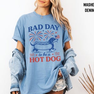 May include: Light blue t-shirt with a graphic of a dachshund hot dog, fireworks, and the text "BAD DAY to be a HOT DOG". The shirt is made of washed denim material.