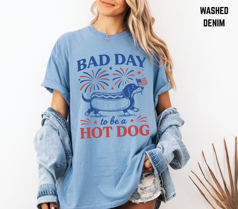 May include: Light blue t-shirt with a graphic of a dachshund hot dog, fireworks, and the text "BAD DAY to be a HOT DOG". The shirt is made of washed denim material.