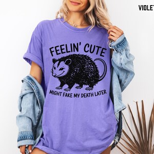 Może przedstawiać: Fioletowa koszulka z napisem "Feelin' Cute" nad czarną ilustracją oposa. Poniżej znajduje się napis "Might Fake My Death Later". Koszulka ma kr&oacute;tkie rękawy i luźny kr&oacute;j.