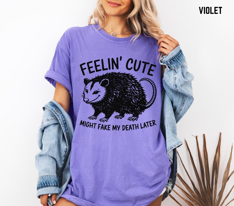 Może przedstawiać: Fioletowa koszulka z napisem "Feelin' Cute" nad czarną ilustracją oposa. Poniżej znajduje się napis "Might Fake My Death Later". Koszulka ma kr&oacute;tkie rękawy i luźny kr&oacute;j.
