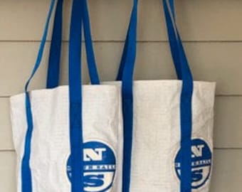 Puede incluir: Dos bolsas de tela blancas con correas azules y un logotipo circular azul con las letras NS y el texto North Sails. Las bolsas están fabricadas con un material texturizado y duradero, y cuentan con asas largas para llevar al hombro.