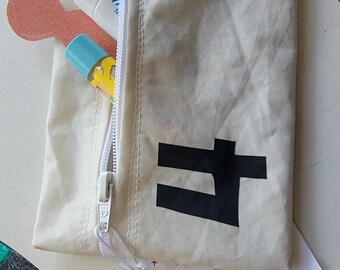 May include: Off-white zipper pouch with a black graphic and a white zipper. The pouch is partially open, revealing a tube of lotion, a small bottle, and a peach-colored item. A tag reads "Zipper Pouch" and the size is 6"W X 5"H.