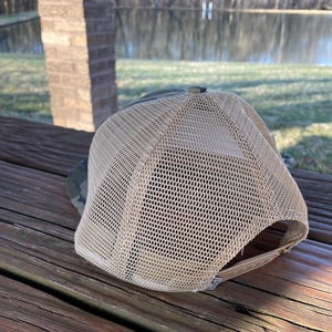 K&ouml;nnte beinhalten: Eine braune und gr&uuml;ne Camouflage-Trucker-Kappe mit einem beigefarbenen Netzr&uuml;cken. Die Kappe liegt auf einer Holzoberfl&auml;che.