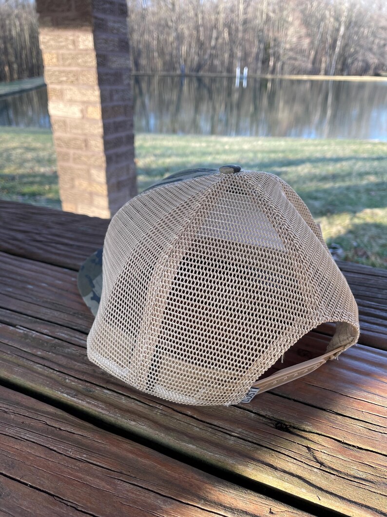 K&ouml;nnte beinhalten: Eine braune und gr&uuml;ne Camouflage-Trucker-Kappe mit einem beigefarbenen Netzr&uuml;cken. Die Kappe liegt auf einer Holzoberfl&auml;che.