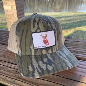 K&ouml;nnte beinhalten: Eine Camouflage-Trucker-Kappe mit braunem Netzr&uuml;cken. Die Kappe hat einen Patch mit wei&szlig;em Hintergrund und einem Bild eines Wei&szlig;wedelhirsches mit Geweih. Der Patch tr&auml;gt auch den Text "Banjo Brand Hat Co."