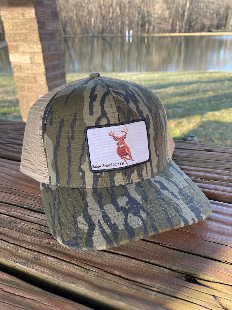 K&ouml;nnte beinhalten: Eine Camouflage-Trucker-Kappe mit braunem Netzr&uuml;cken. Die Kappe hat einen Patch mit wei&szlig;em Hintergrund und einem Bild eines Wei&szlig;wedelhirsches mit Geweih. Der Patch tr&auml;gt auch den Text "Banjo Brand Hat Co."