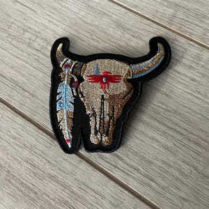 Puede incluir: Parche bordado con un cráneo de toro con cuernos, una pluma y detalles de inspiración nativa americana. El cráneo es de color beige y marrón, con detalles en rojo y azul. El parche tiene un borde negro.