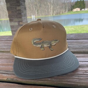 May include: A brown and green baseball cap with an embroidered alligator on the front. The cap has a white rope trim around the brim.
