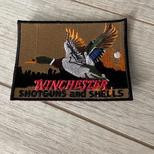 May include: Embroidered patch with a flying duck design, set against a brown backdrop with a sunset. The patch features the text "Winchester Shotguns and Shells" in red and black.