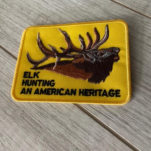 May include: Embroidered patch with a yellow background and a brown elk head with large antlers. The text "ELK HUNTING AN AMERICAN HERITAGE" is displayed in black letters. The patch has a yellow border.