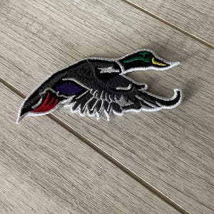 May include: Embroidered patch featuring a detailed mallard duck in flight. The patch showcases a black body with accents of green, yellow, purple, red, and white. The duck's wings are spread, and the design is outlined in white.