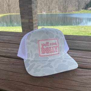 May include: White camouflage trucker hat with a pink patch that says "y'all need dolly".