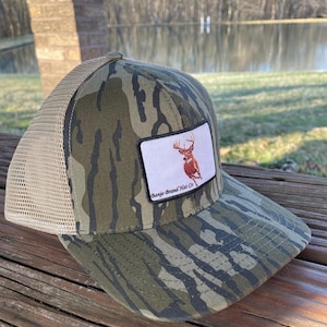 K&ouml;nnte beinhalten: Eine Camouflage-Trucker-Kappe mit einem braunen Hirsch-Patch und dem Text "Bango Brand Hat Co."