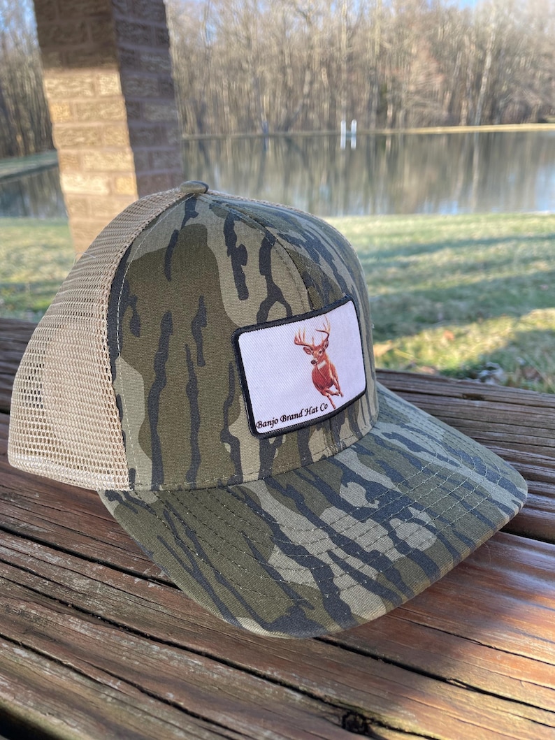 K&ouml;nnte beinhalten: Eine Camouflage-Trucker-Kappe mit einem braunen Hirsch-Patch und dem Text "Bango Brand Hat Co."