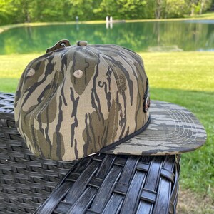 May include: A camouflage baseball cap with a flat peak. The cap has a brown, green, and tan pattern. The cap is resting on a dark wicker chair. The background includes a pond and trees.