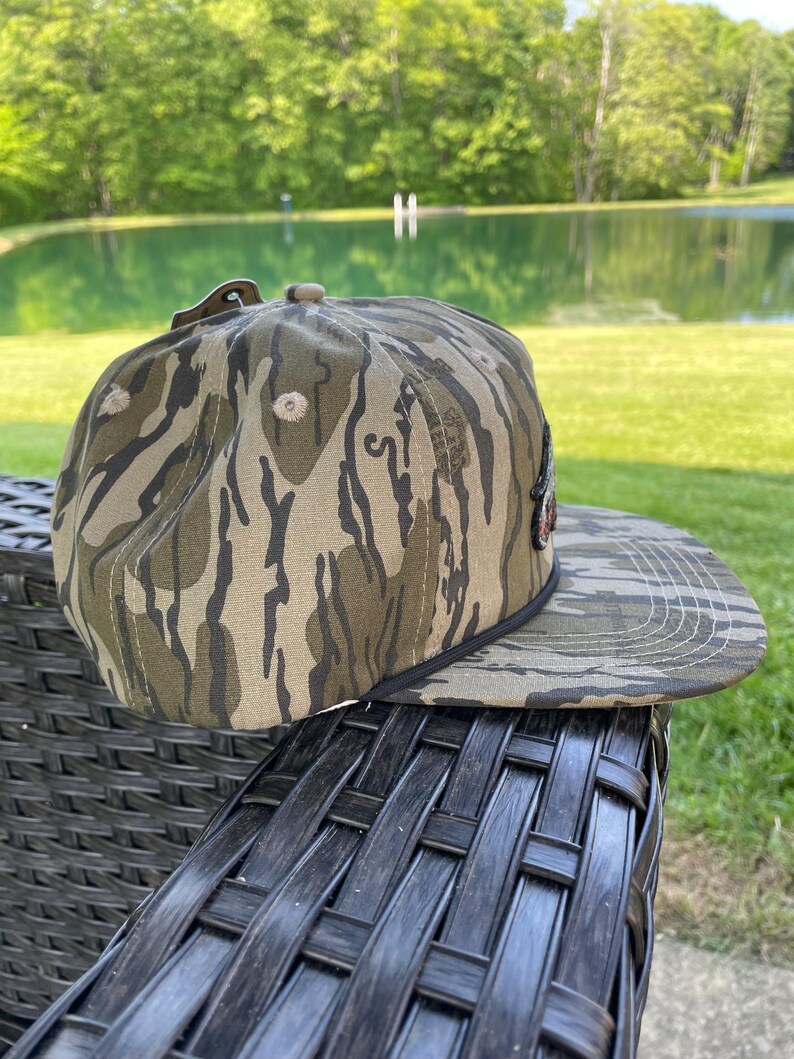 May include: A camouflage baseball cap with a flat peak. The cap has a brown, green, and tan pattern. The cap is resting on a dark wicker chair. The background includes a pond and trees.