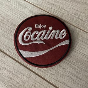 May include: A circular embroidered patch with a burgundy background and a black border. The patch features the word "Enjoy" above the word "Cocaine" in a white, stylized font, with a white swoosh design.