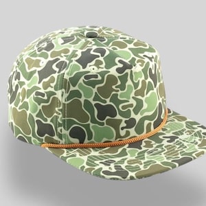 May include: A green and brown camouflage patterned baseball cap with a brown rope trim around the brim.