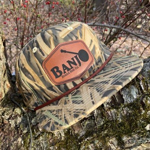 Puede incluir: Una gorra de béisbol de camuflaje con un parche de cuero marrón que dice "BANJO Brand Hat Co."