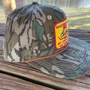 May include: A camouflage baseball cap with a brown leather band and a yellow patch that reads "Banjo Brand Music Co. Est. Eat More Catfish".
